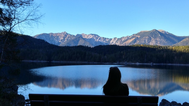 Zdjęcie z tyłu widok sylwetki kobiety siedzącej na ławce nad jeziorem.
