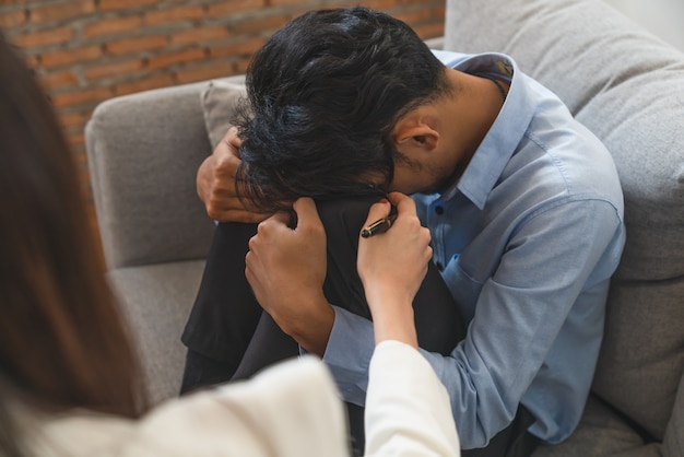 Zaakcentowany Azjatycki Młody Człowiek Cierpi Na Problem Z życiem, Siedząc Na Kanapie Z Rękami Trzymając Głowę, Podczas Gdy Kobieta Psychiatra Próbuje Go Pocieszyć