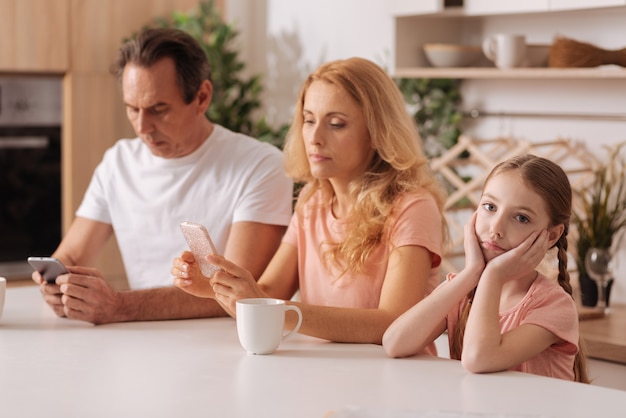 Zaangażowana obsesja uzależnionych rodziców siedzących w domu i korzystających z elektronicznych gadżetów, podczas gdy mała córka wyrażająca smutek i czekająca na rodziców
