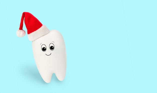Zabawka biały ząb w czerwonym czapce Świętego Mikołaja na białym tle na jasnoniebieskim tle. Świąteczna koncepcja kliniki stomatologicznej. Świąteczna i noworoczna medyczna karta zimowa, ładny charakter plakatu, miejsce na kopię.