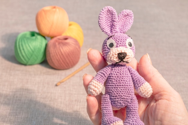 Zabawka króliczek w kobiecej dłoni na tle dziewiarskich piłek. Hobby zabawki szydełkowe.