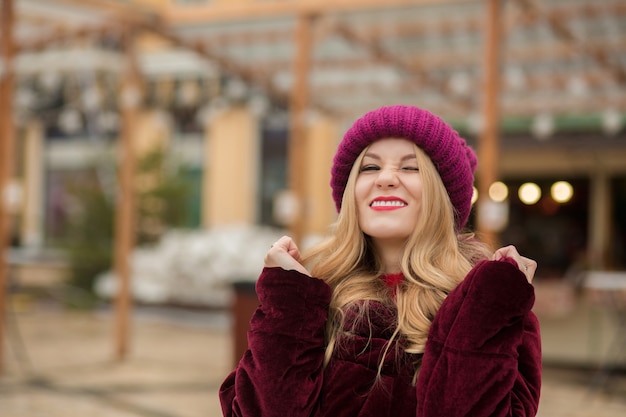 Zabawna blondynka w czerwonej czapce i płaszczu zimowym, pozuje na tle girlandy na ulicy w Kijowie