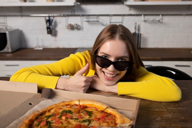 Zabawna brunetka dziewczyna w żółtym swetrze i czarnych okularach jedzenie pizzy w kuchni