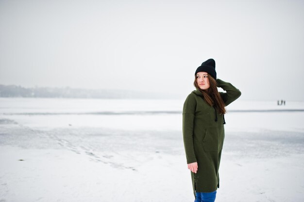Zabawna dziewczyna nosi długie zielone dżinsy i czarne nakrycia głowy nad zamarzniętym jeziorem w zimowy dzień