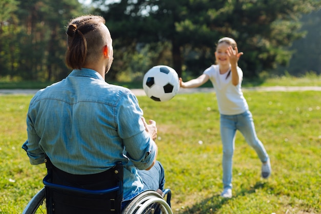 Zabawna gra. Widok młodego mężczyzny na wózku inwalidzkim z tyłu, łapiącego piłkę rzuconą przez jego małą dziewczynkę i cieszącego się weekendem w parku