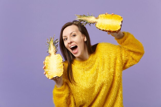 Zabawna młoda kobieta w futro sweter utrzymując otwarte usta trzymając połówki świeżych dojrzałych owoców ananasa na białym tle na fioletowym tle pastel. Ludzie żywy styl życia zrelaksować koncepcję wakacji. Makieta miejsca na kopię.