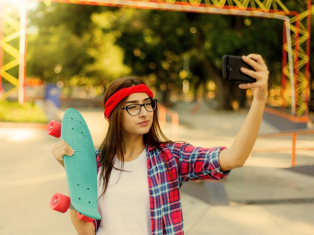 Zabawna młoda kobieta w okularach i czerwoną koszulę w kratę. Hipster dziewczyna n skatepark, a sprawia, że selfie z deskorolką w skateparku w słoneczny jasny dzień. Letnia zabawa