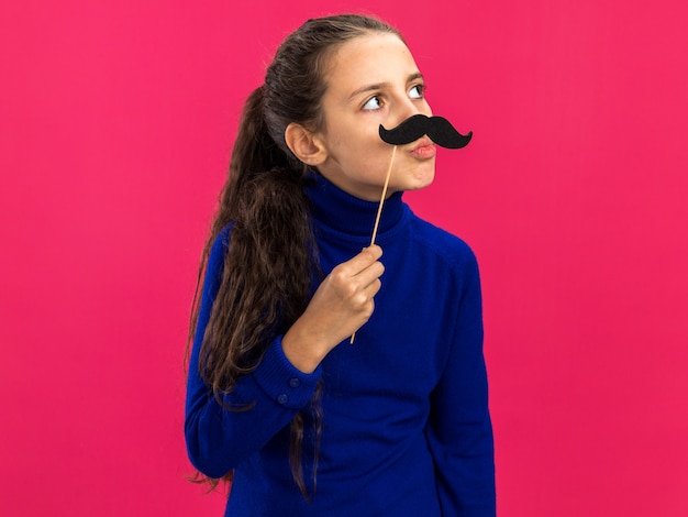 Zabawna Nastolatka Trzyma Fałszywe Wąsy Na Patyku Nad Ustami, Patrząc Na Bok Z Zaciśniętymi Ustami Na Różowej ścianie Z Miejsca Kopiowania
