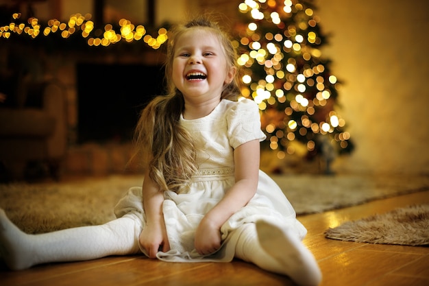 Zabawna roześmiana dziewczyna ubrana w białą sukienkę pozuje na podłodze w bożonarodzeniowym wnętrzu z choinką