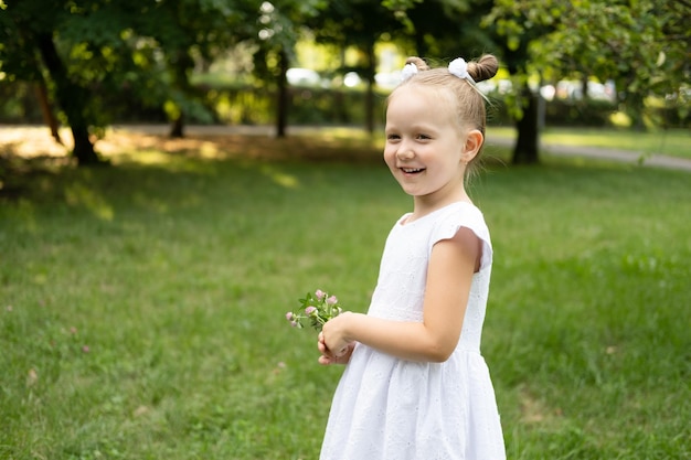 zabawna uśmiechnięta dziewczynka w białej sukience z wiosennymi kwiatami w zielonym parku