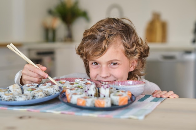 Zabawne dziecko z kręconymi włosami uśmiecha się i patrzy na kamerę, jedząc rolki sushi pałeczkami i chowając się za talerzami w kuchni w domu