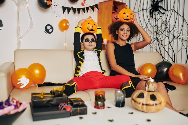 Zabawny brat i siostra z makijażem Halloween siedzą na kanapie w urządzonym pokoju i pozują z lampionami Jack-o na głowach