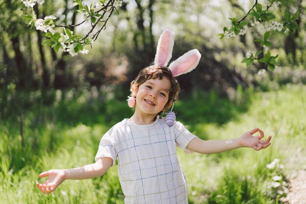 Zabawny chłopiec z koszykiem jaj i uszami królika na polowaniu na pisanki w słonecznym wiosennym ogrodzie