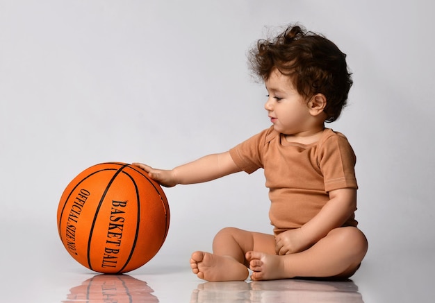 Zabawny, kręcony mały chłopiec ubrany w body siedzi boso i gra w nową koszykówkę na szarym tle Miejsce na tekst Pojęcie aktywnego dzieciństwa