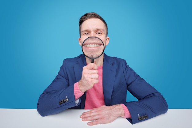 Zabawny młody człowiek trzyma szkło powiększające jego uśmiech toothy, siedząc przy biurku w izolacji