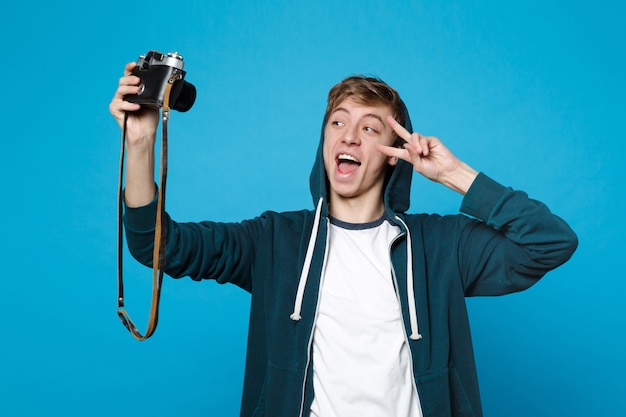 Zabawny młody człowiek w ubranie robi selfie strzał na retro vintage aparat fotograficzny pokazując znak zwycięstwa na białym tle na niebieskiej ścianie. Ludzie szczere emocje, koncepcja stylu życia.