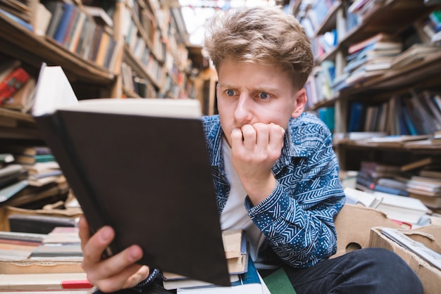 Zabawny student siedzący na podłodze w starej bibliotece publicznej i czytający książkę z zainteresowanym spojrzeniem.