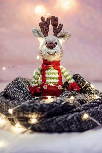 Zabawny świąteczny jeleń w swetrze w paski i stylowy czerwony motylkowy krawat w pobliżu lampek choinkowych na różu.