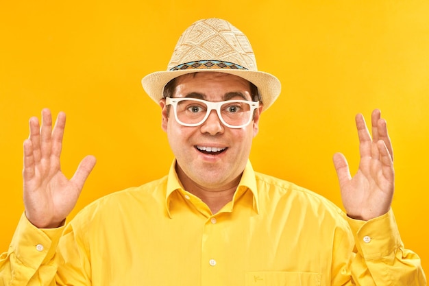 Zabawny turysta grubas w kapeluszu i okularach z zaskoczoną miną rozkłada ręce na żółtym tle studia