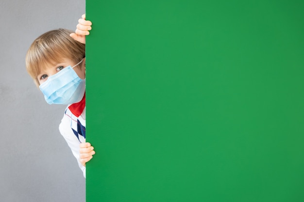 Zabawny uczeń dziecko noszenie maski ochronnej w klasie. Szczęśliwe dziecko chowając się za zieloną tablicą.