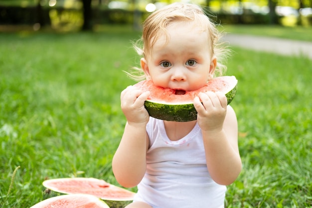 zabawny uśmiechnięty chłopiec w białym body jedzący arbuza na zielonym trawniku