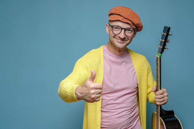 Zabawny uśmiechnięty mężczyzna w jasnych ubraniach trzyma w dłoni gitarę akustyczną i pokazuje gest aprobaty