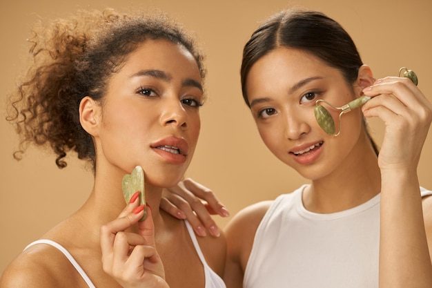 Zabiegi kosmetyczne atrakcyjne młode kobiety patrzące na kamery za pomocą jadeitowego wałka i twarzy gua sha