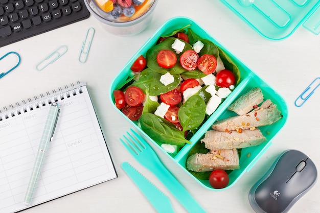 Zabierz pudełko na lunch ze świeżą sałatką i tuńczykiem na biurko z artykułami biurowymi.