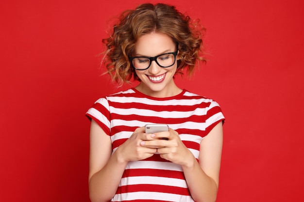 Zachwycona młoda kobieta w pasiastej koszulce i modnych okularach czytająca nieoczekiwaną wiadomość na smartfonie z uśmiechem na czerwonym tle