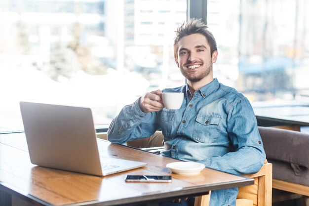 Zachwycony uśmiechnięty szczęśliwy młody człowiek freelancer pracujący na laptopie po przerwie pijącej kawę