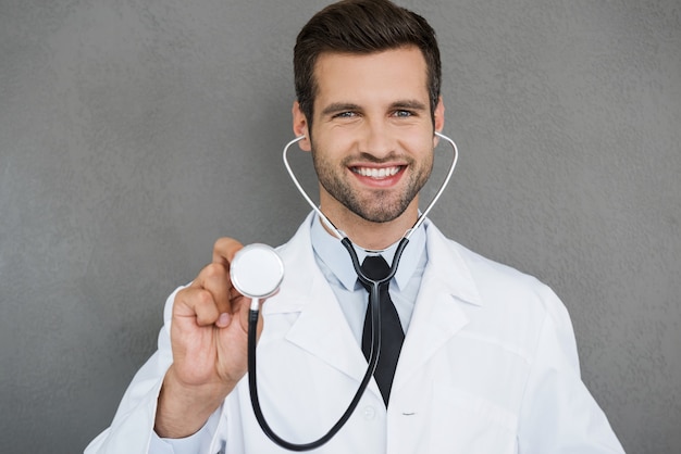 Zacznę egzamin! Uśmiechnięty młody lekarz w białym mundurze, noszący stetoskop i patrzący na kamerę, stojąc na szarym tle
