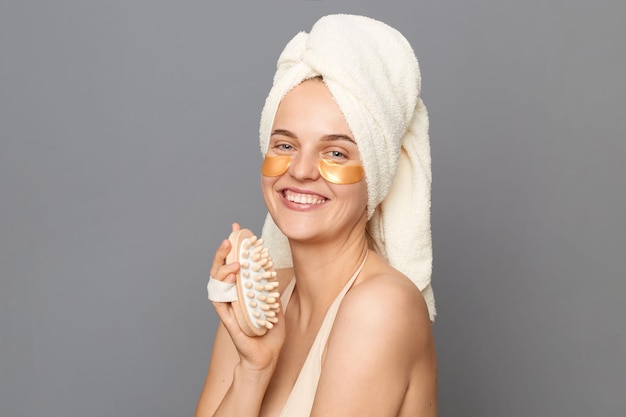 Zadbana, ładna młoda kobieta owinięta ręcznikiem za pomocą szczotki do ciała do masażu na sucho stojące plastry pod oczami, pozując na białym tle na szarym tle puste miejsce na tekst promocyjny