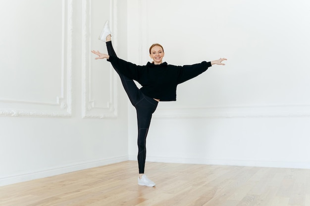 Zadowolona baletnica balansuje na jednej nodze, rozciąga się w studio, ćwiczy jogę, wykazuje dobrą elastyczność, ubrana na czarno, ma radosny wyraz twarzy, ćwiczy w pomieszczeniu.