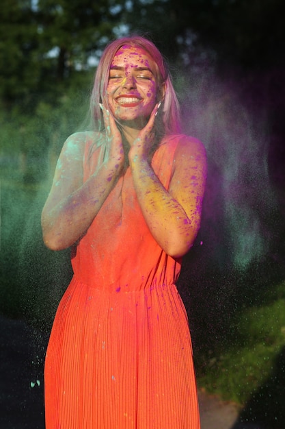 Zadowolona blond modelka w pomarańczowej sukience pozuje w chmurze suchej farby na festiwalu Holi