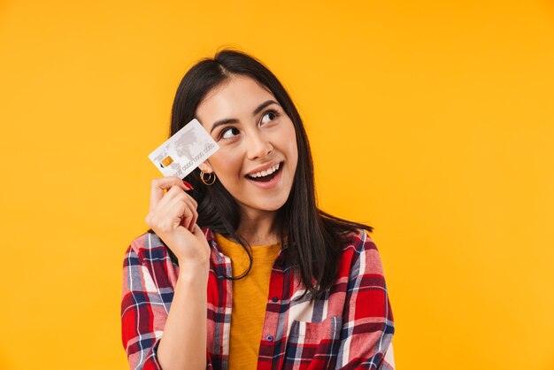 zadowolona brunetka uśmiecha się trzymając kartę kredytową odizolowaną nad żółtą ścianą