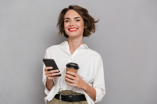zadowolona kobieta trzyma kawę na wynos i przy użyciu telefonu komórkowego, odizolowane na szarej ścianie