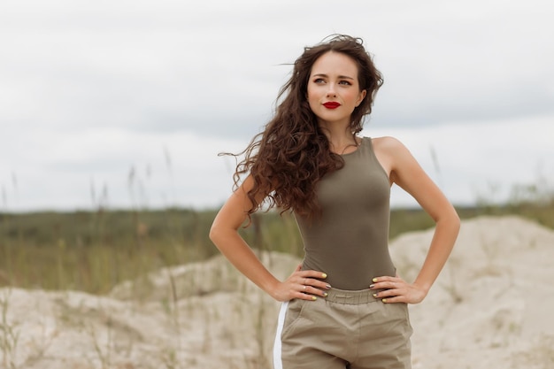 Zadowolona kobieta w modnym stroju, z czerwonymi ustami, stojąca na plaży z rękami w pasie