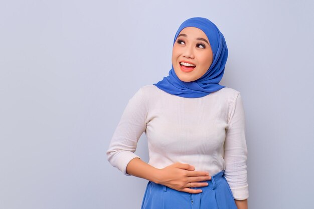 Zadowolona młoda azjatycka muzułmanka trzyma obie ręce na brzuchu po pysznej kolacji odizolowanej na białym tle