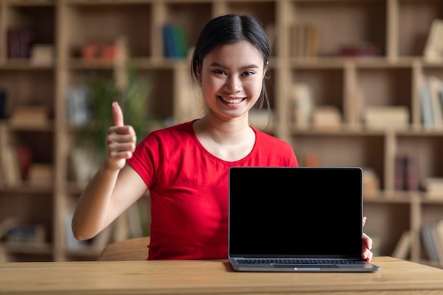 Zadowolona młoda japońska uczennica z zespołem Downa pokazuje kciuk w górę i laptop z pustym ekranem, który zatwierdza edukację