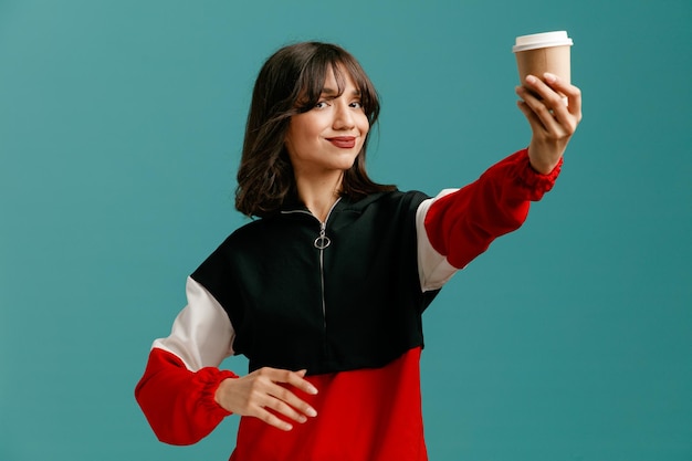 Zadowolona młoda kobieta kaukaski trzymając rękę w powietrzu, patrząc na aparat rozciągający filiżankę kawy na wynos w kierunku aparatu na białym tle na niebieskim tle