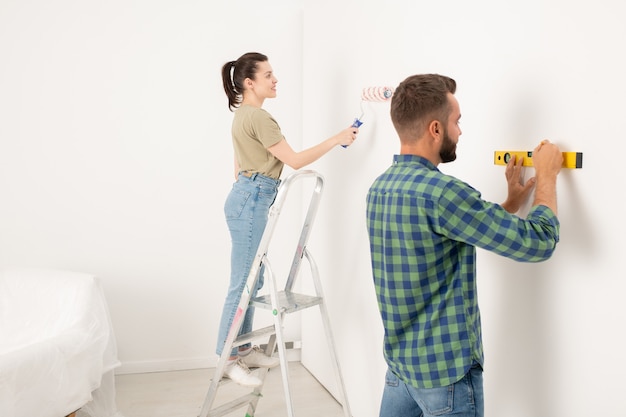 Zadowolona młoda kobieta stojąca na drabinie i malująca ścianę, podczas gdy jej chłopak rysuje linię za pomocą poziomu