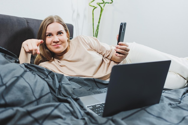 Zadowolona młoda kobieta z telefonem kontynuująca pracę na laptopie Kobieta siedząca z komputerem w łóżku, studiująca w domu nauczanie na odległość i koncepcja pracy zdalnej