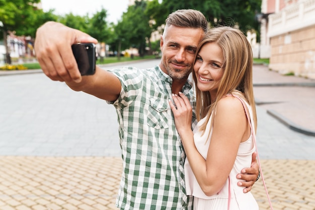 zadowolona młoda para w letnich ubraniach uśmiecha się i robi wspólne zdjęcie selfie podczas spaceru ulicą miasta