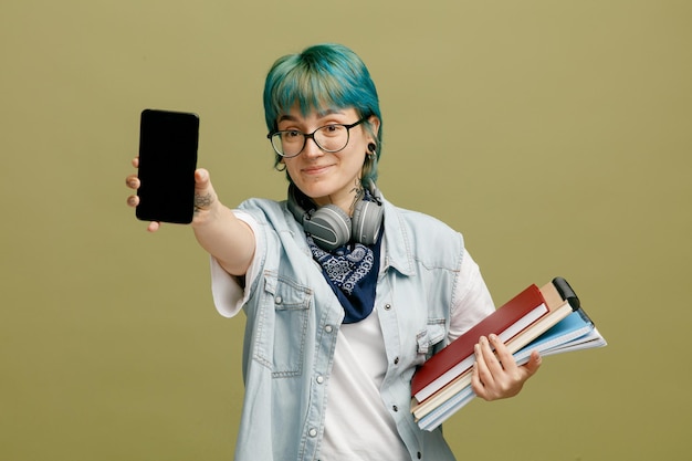 Zadowolona Młoda Studentka W Okularach Bandana I Słuchawki Na Szyi Trzymająca Notesy Patrząc Na Aparat Rozciągający Telefon Komórkowy W Kierunku Kamery Na Białym Tle Na Oliwkowo-zielonym Tle