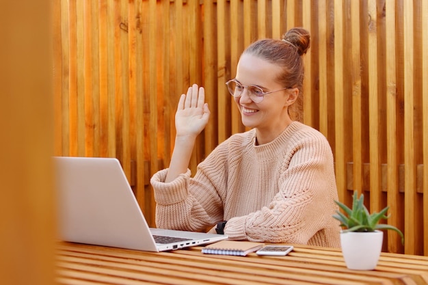 Zadowolona piękna kobieta z fryzurą kok, ubrana w beżowy sweter i okulary, pracuje na laptopie i prowadzi rozmowę wideo online, machając ręką do ekranu, pozując na drewnianej ścianie