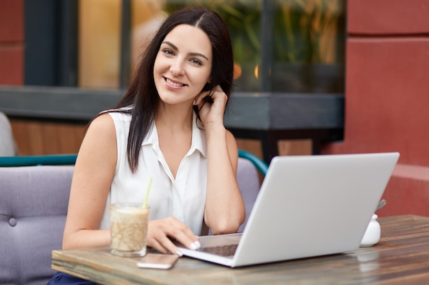 Zadowolona, przyjemnie wyglądająca freelancerka wygląda z dystansem na notebooka, pije koktajl mleczny, pozytywnie patrzy w kamerę, lubi swoją pracę, pozuje w restauracji na świeżym powietrzu, łączy się z szybkim Internetem