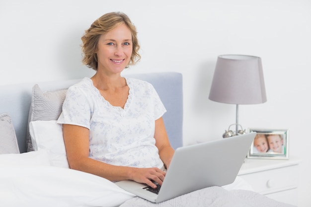 Zadowolony blondynki kobiety obsiadanie w łóżkowym używa laptopie