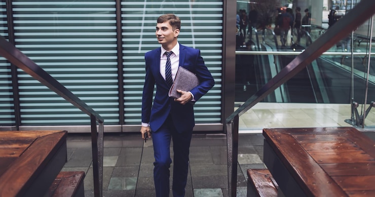 Zadowolony elegancki przystojny młody biznesmen w drogim kostiumie wspina się po schodach w centrum biznesowym z szarym folderem i odwraca wzrok na tle miejskim