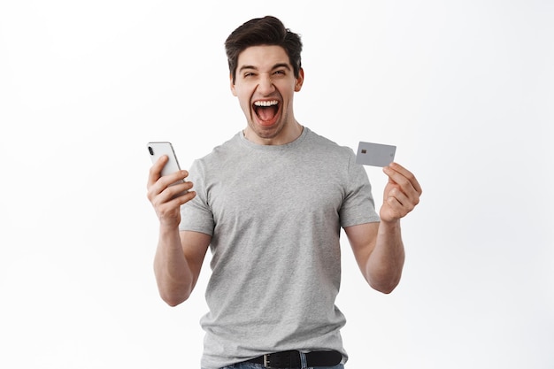 Zadowolony facet świętuje, pokazuje plastikową kartę kredytową i telefon komórkowy, krzyczy zdumiony, triumfuje, wygrywa pieniądze, stojąc na białym tle