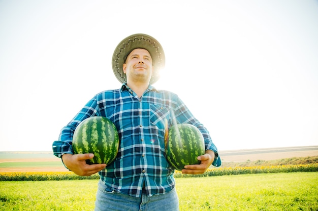 Zadowolony farmer z dwoma arbuzami na polu w słoneczny letni dzień mężczyzna w kapeluszu na głowie...